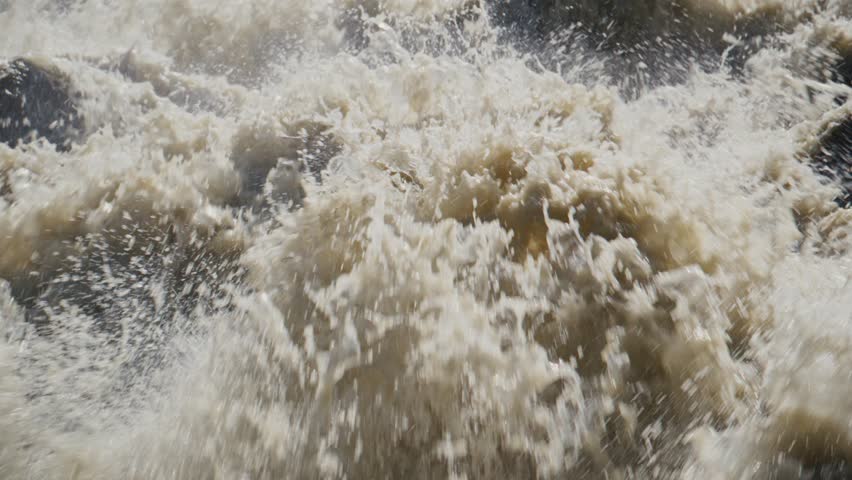 Raging river water crashes with force. White foam explodes against dark rocks. Turbulent flow shows raw nature's power. Ideal for adventure, environmental, or extreme sports visuals
