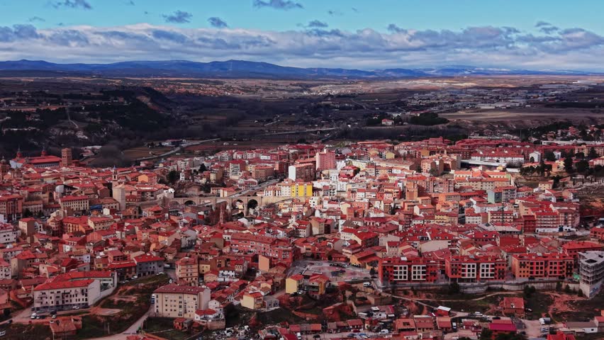A small town in Spain shows buildings with red roofs. The view includes hills and fields in the distance. The sky has clouds on a clear day. People go about their daily activities.