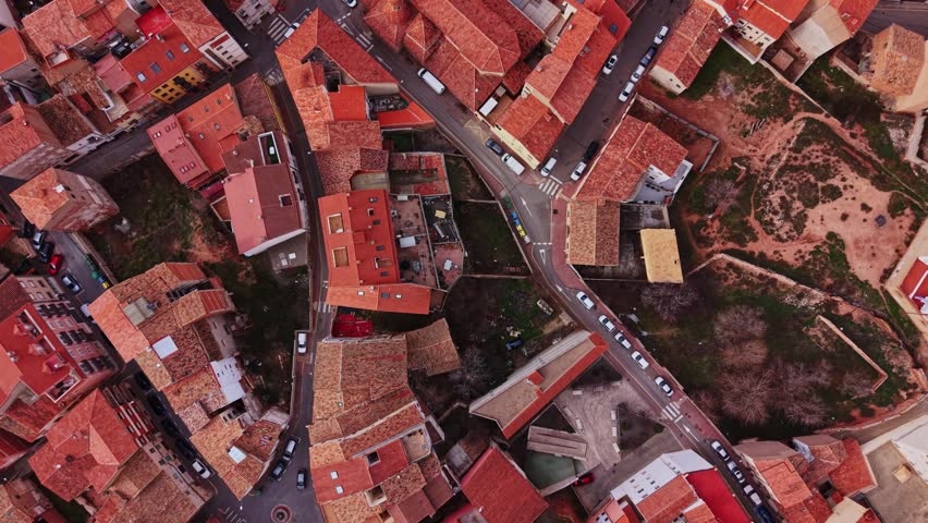 Bird's eye view shows the red roofs and narrow streets of a small town in Spain. Cars are parked along the roads, and green spaces are nearby. The scene shows daily life in the town.