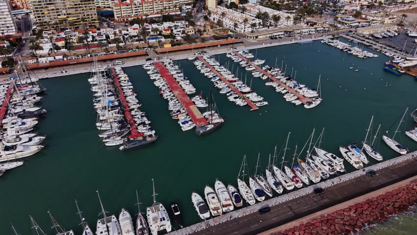 Many boats and yachts are docked in a marina in Spain. People move about the docks. The sun shines above, and buildings are seen in the background.