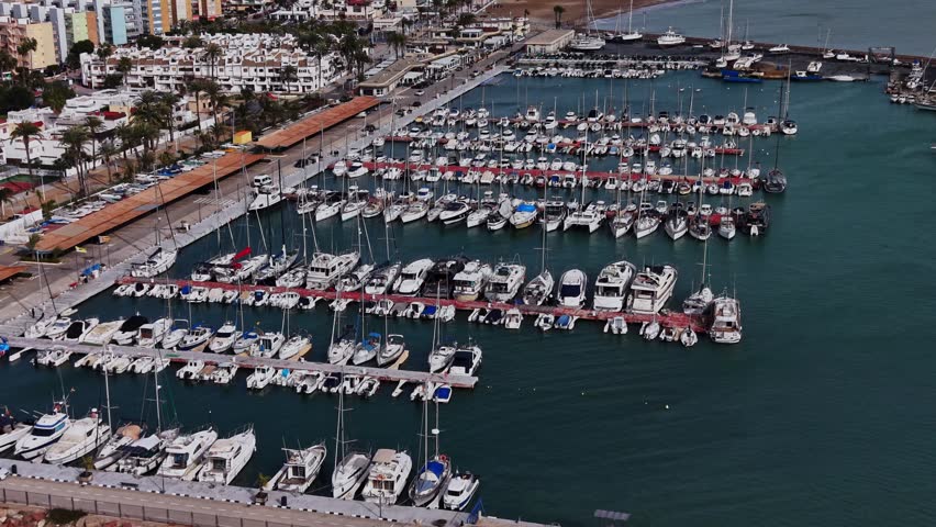 Many boats are docked in a marina in Spain. The marina is busy with several yachts and smaller boats. Buildings can be seen in the background along the waterfront.