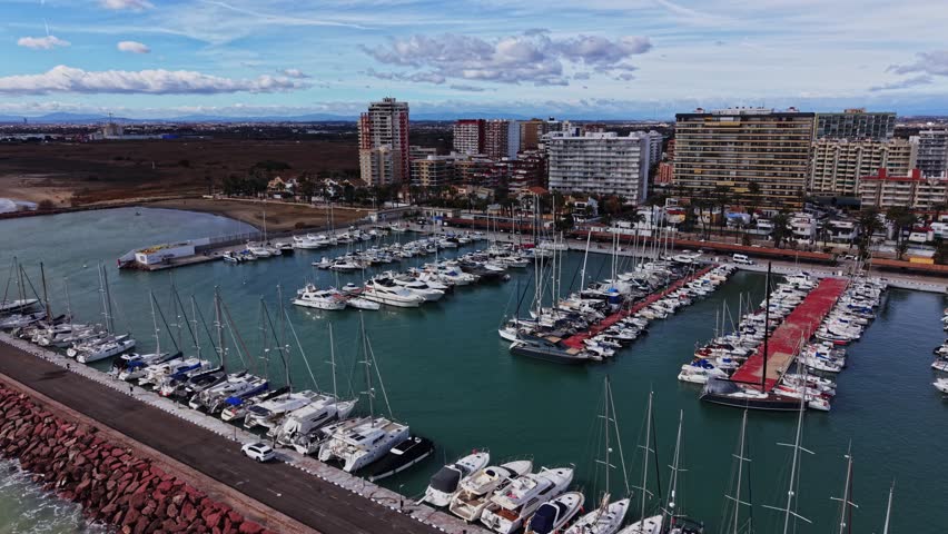 People enjoy their time at the marina in Spain. Boats are docked in the water, and buildings are visible in the background. The sky shows clouds and a hint of blue.
