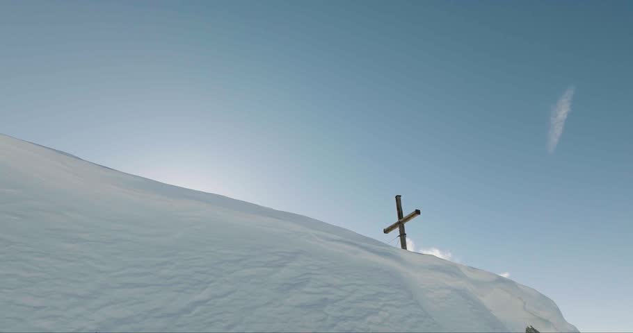 Aerial view of a wooden cross on a snow-covered mountain peak, with snow-capped mountains in the background under a blue sky, Arlberg, Austria.