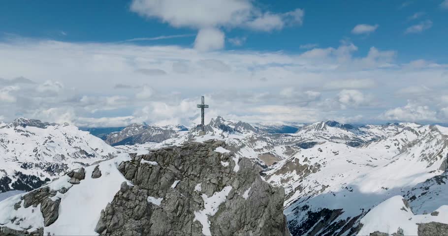 Aerial view of a stark, snow-covered mountain peak crowned with a cross, contrasting with the majestic, snow-capped peaks in the distance, Arlberg, Austria.