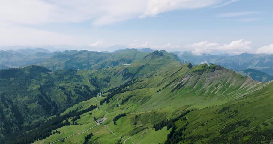 Aerial view of rolling green mountains, textured slopes, and distant peaks under a blue sky with scattered clouds, Wandern, Austria.
