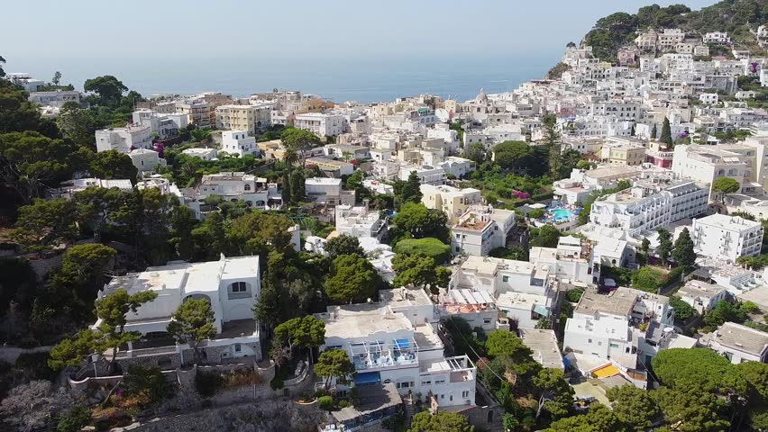 An aerial view showcases the hillside town of Capri, Italy, with white buildings nestled among green trees, overlooking the blue Mediterranean Sea.