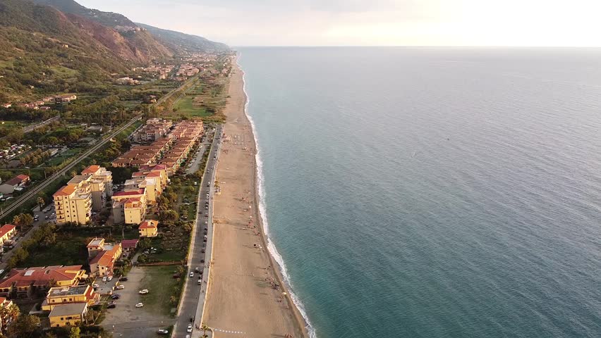 An aerial view captures the long sandy beach and coastal town along the Tyrrhenian Sea in Calabria, Italy. Mountains rise in the background.