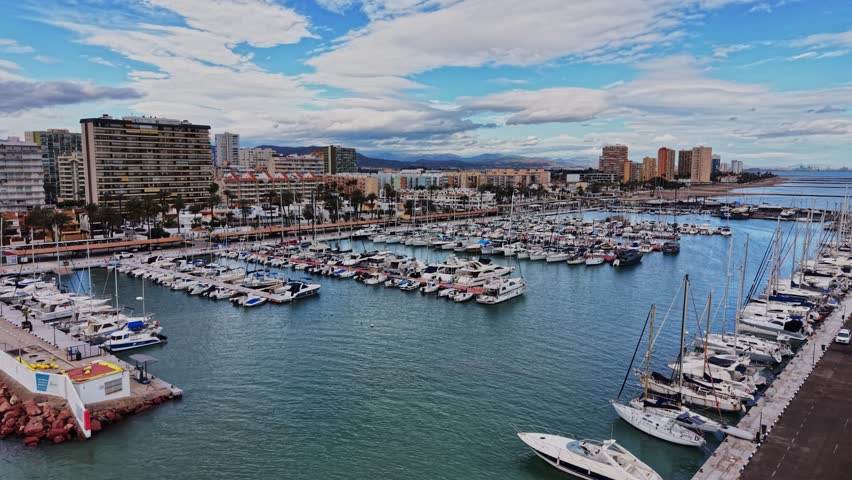 Many boats are docked in the harbor in Spain. Buildings line the coast, and the sky is partly cloudy. People may explore the area around the water in this lively location.