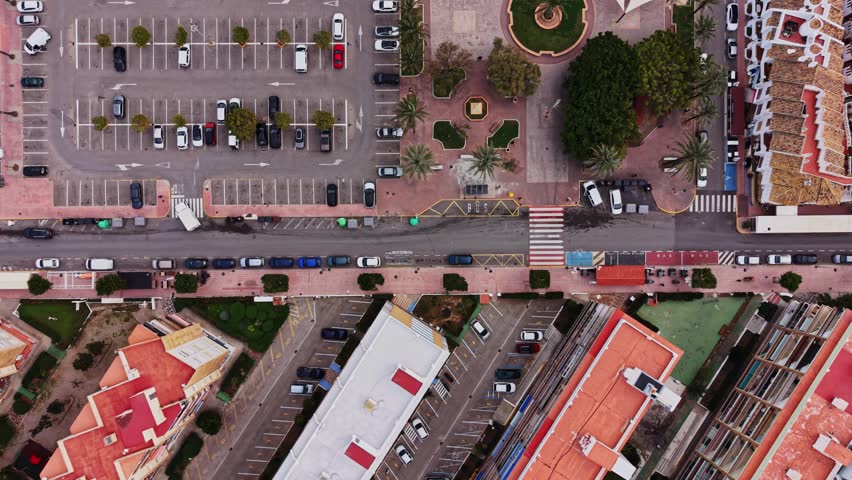Aerial view of a busy street in Spain showing parked cars and buildings. The scene includes palm trees and a square with a fountain. People walk through the street and parking lot.