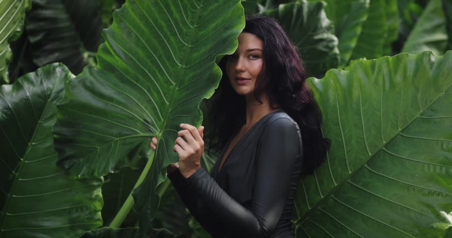 Caucasian woman with glowing skin in a black outfit posing peacefully in lush tropical foliage. Conceptual beauty, skincare, fashion, and nature promo in high quality footage. 