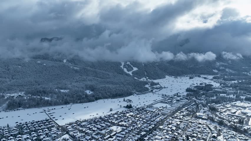 Aerial view of a snow-laden landscape with a town nestled against a backdrop of mountains shrouded in clouds, Garmisch-Partenkirchen, Bavaria, Germany.