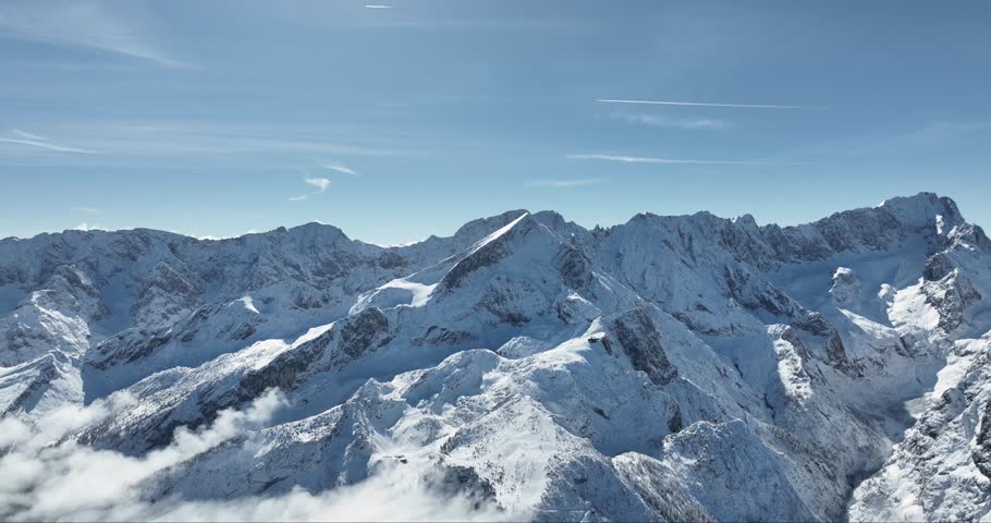 Aerial view of snow-capped mountains piercing through the clouds under a clear blue sky, creating a striking contrast in Garmisch-Partenkirchen, Bavaria, Germany.