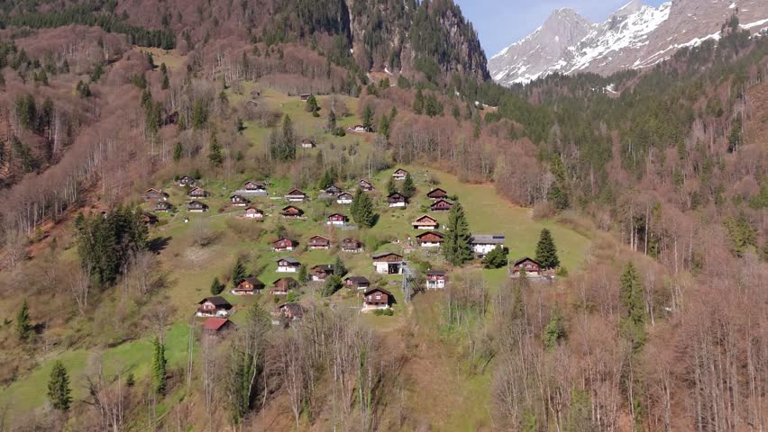 Alpine hamlet of Schlattberg, Luchsingen, Switzerland with traditional chalets across a steep hillside surrounded by forest and snow-capped mountains. Aerial drone shot.