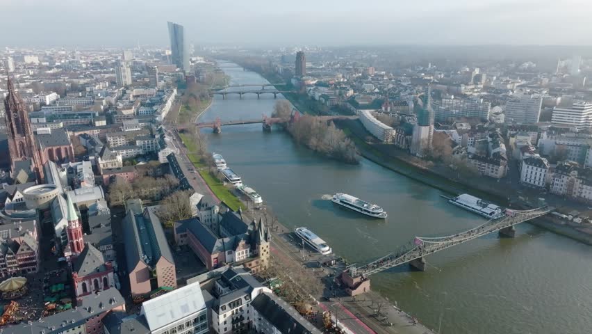Aerial view of Frankfurt: Main river with bridges and boats, Frankfurt Cathedral and old town on the left, modern skyline in the background under a clear sky.