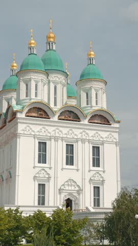 Majestic white Orthodox cathedral stands against muted sky. Green domes topped with golden crosses create striking contrast. Intricate facade details treetops visible below. Serene, historic mood