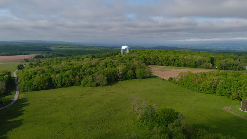 Aerial drone view of white water tower rising above dense forest on Chestnut Ridge near Morgantown, surrounded by rolling hills and greenery under partly cloudy sky
