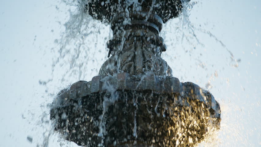 Close-up view of an ornate tiered fountain with water splashing and cascading over decorative bronze sculpture. Golden sunlight illuminates droplets against a pale sky background.