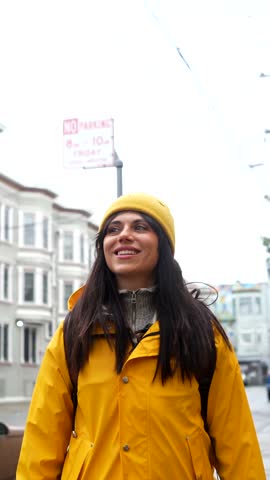 Joyful young woman wearing a yellow raincoat and beanie smiling while exploring the city streets of san francisco, enjoying her vacation and sightseeing adventure in an urban environment