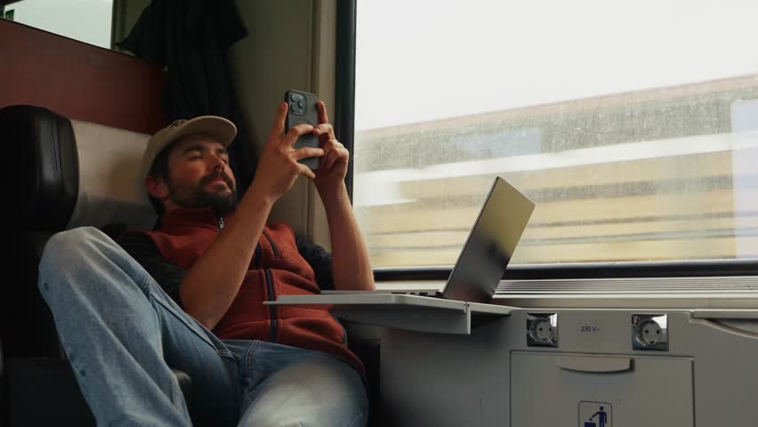 A Man Relaxes on a Train, Using His Phone With a Laptop on the Table - Medium Shot