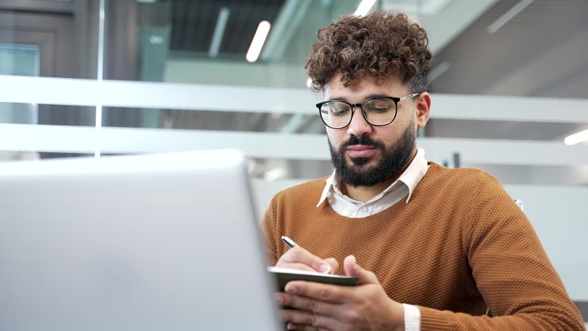 Businessman watching video call conference or training takes notes in a notebook looking at laptop screen sitting in business office. Worker listening to remote online course, seminar. Close up