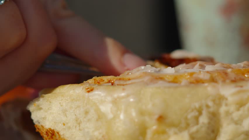 Close-up of Fresh Soft Delicious Cinnamon Bun with Female Hand Using Fork to Cut in Background. Outdoor Al Fresco Dining. Sweet Dessert Bun with Icing Glaze.
