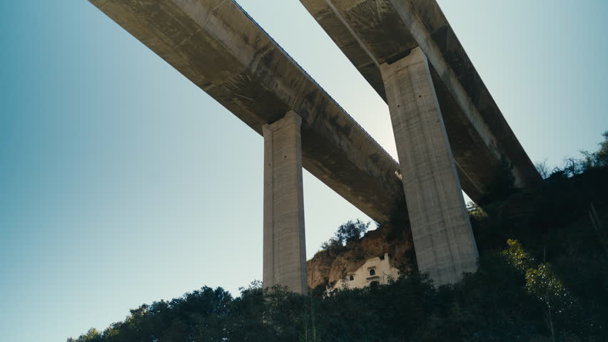 Low angle view of a large concrete viaduct spanning a canyon with towering pillars and a small house beneath. Strong contrast between infrastructure and natural landscape under clear blue sky.