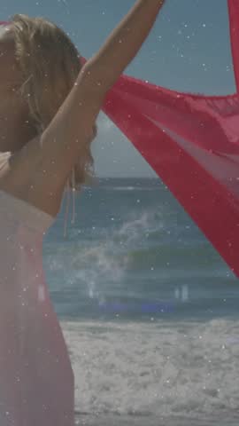 Vertical video: Woman raising arms and unfurling red fabric on beach for fashion visuals. Scarf, coastline, ocean, breeze, horizon, dress, vertical video