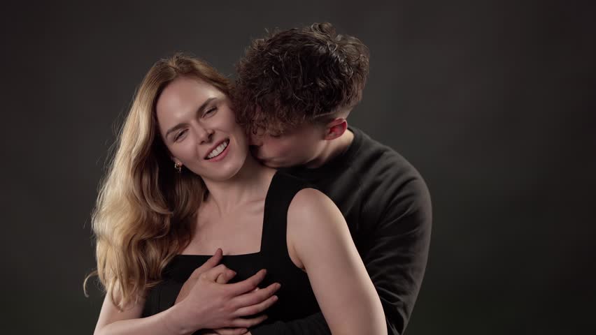 Young man kissing his girlfriend on the neck while they embrace against a dark background