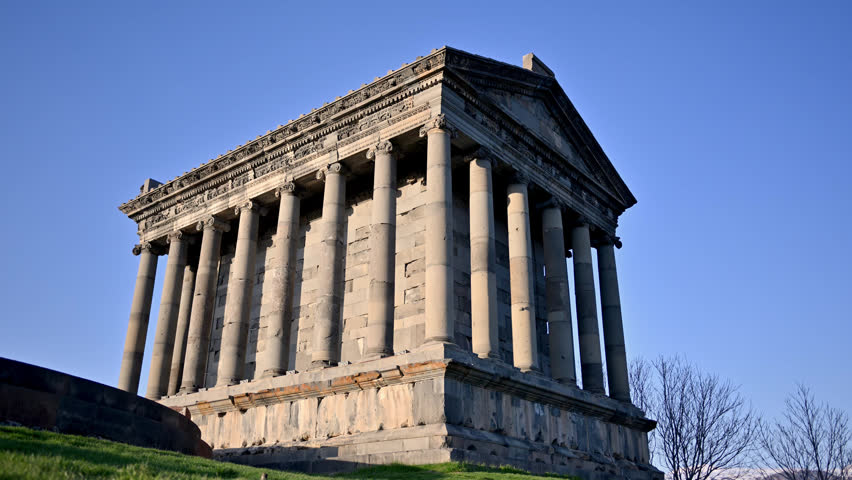 Ancient stone Garni temple stands tall on grassy hill. Clear blue sky above historic structure. Bare trees frame sides of monument. Stone columns support triangular roof. Structure shows weathered texture