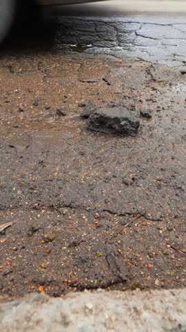Vertical close-up of broken asphalt after winter with loose chunks and a passing car wheel, showing road wear, pothole damage, and seasonal street deterioration.