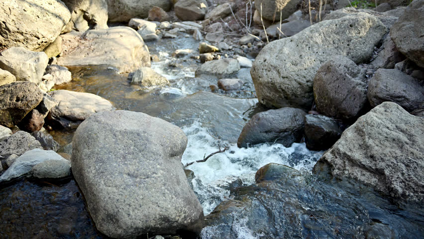 Small stream flows over smooth gray stones. Water splashes around boulders. Clear water moves fast through rocky terrain. Natural forest environment surrounds the creek