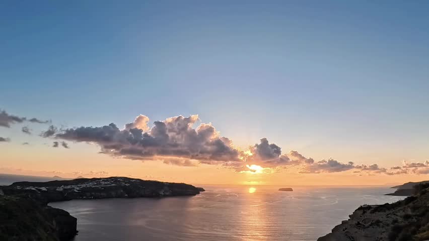 Stunning time-lapse of a golden sunset over the volcanic caldera of Santorini, Greece. The video shows the sun disappearing behind clouds and the horizon, reflecting on the Aegean Sea with a vibrant orange and pink sky.