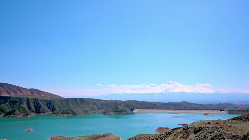 Vast turquoise lake nestled between arid hills and distant mountains. Clear blue sky stretches above calm waters