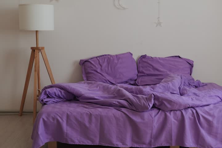 A bed with purple sheets is unmade, pillows are scattered. A tall lamp stands nearby, providing light in the simple room during the day