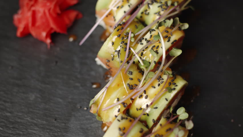 Delicious sushi uramaki roll with avocado, tuna, and salmon rotating on a dark slate plate, showing traditional Japanese food, cuisine, and fine dining concept