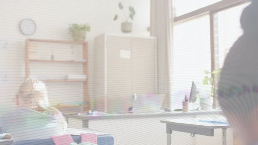 Teacher walking toward desks, girl with high bun turning and raising hand to answer in classroom. Instructor, students, participation, engagement, learning, education, sunlit