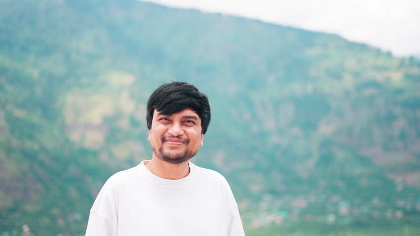 4K Portrait of an Indian male vlogger removing fly insect in front of his face while standing in front of the Himalayan mountain peaks during the summer season in Manali, Himachal Pradesh, India.