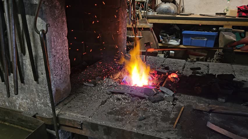 A rustic blacksmith forge glows with bright flames and red embers in a cluttered metalworking workshop, surrounded by coal, tools and workbenches that convey heat, craftsmanship and hands-on tradition.