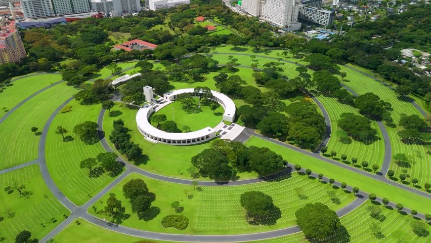 Drone parallax over Manila American Cemetery revealing circular white memorial rotunda surrounded by rows of grave markers on vast green lawns
