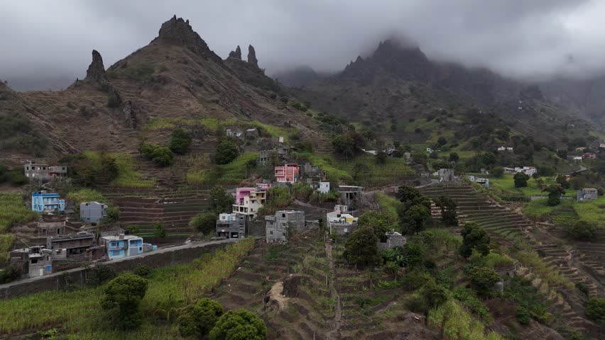 Aerial drone view of a mountain village in Santo Antao, Cape Verde. Scenic terraced fields and colorful houses nestled against jagged volcanic peaks under a misty, dramatic sky