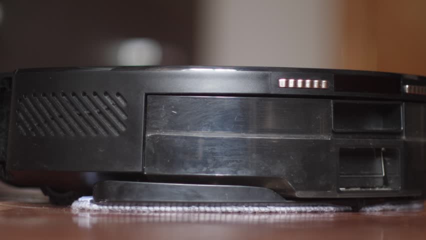 A black robot vacuum cleaner rotates in place and begins moving across a wooden floor. Side view of the sensors working and the automatic cleaner moving in the apartment