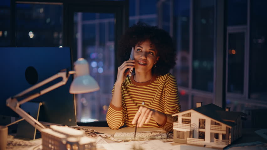 Real estate manager calling cellphone sitting evening workplace closeup. Smiling woman property agent consulting by smartphone in office. African american designer talking phone holding house model.