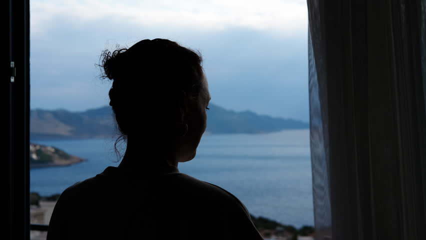 Silhouette of a woman closing curtains in a hotel room with a sea view. Silhouette of a lonely woman looking at the seascape from her hotel room window and then closing the transparent curtains