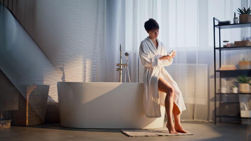 Woman putting legs cream indoors. Relaxed brunette lady preparing foam bathtub sitting at morning sunshine interior. Serene white gown model resting weekend atmosphere having skincare routine alone 