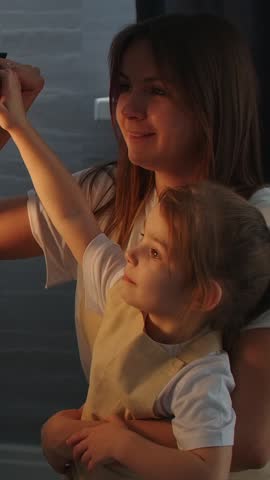 Smiling mother gently holding her young daughter in warm evening light as they reach up together, sharing a tender family moment in a cozy home interior