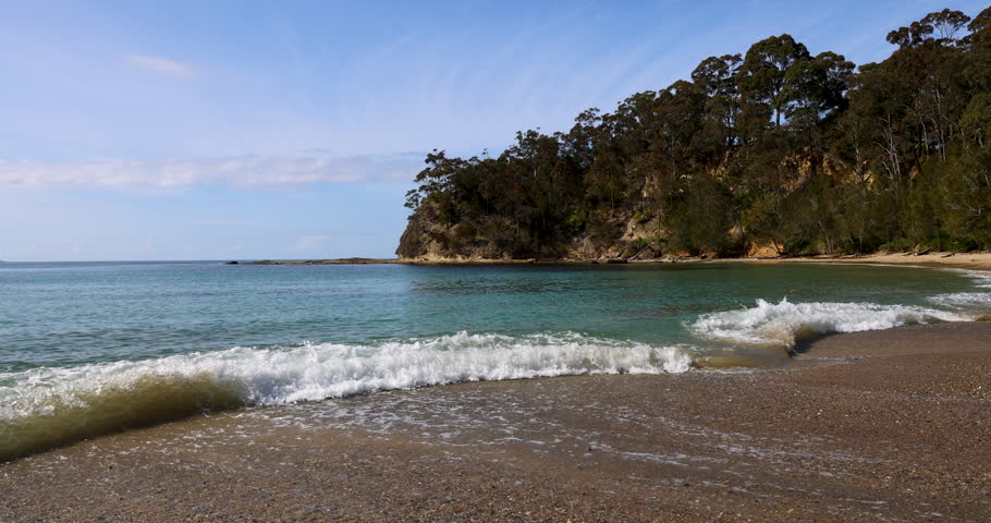 A beautiful coastal cove with waves washing a long a pebbled shoreline under a blue sunny sky. Captured in slow motion from a low perspective.