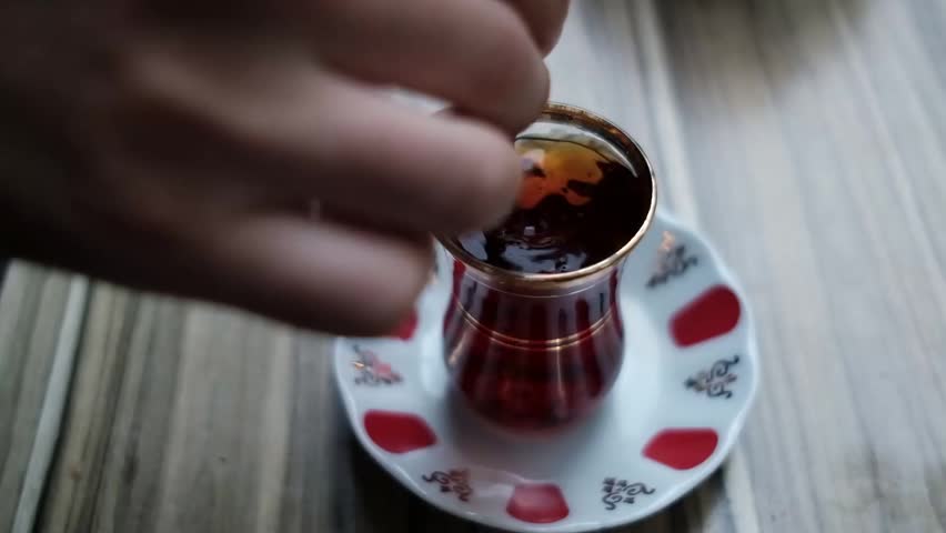 A hand stirs dark tea or coffee inside traditional glass cup, with gold rim. Beverage rests on white ceramic saucer adorned with red circular motifs, small black floral designs. Wooden slat table, neutral background, provides warm natural setting. Soft light creates shimmering reflection along gold rim, highlighting rich deep color liquid. Hand uses spoon, mixing drink before lifting away. Surface remains calm, few bubbles. Footage captures quiet relaxation, hospitality. Perfect for morning routines, breakfast, slow living concepts, cozy scenes. Ideal for culinary lifestyle, traditional tea ceremonies, or morning atmospheres. Cinematic shot, intimate.