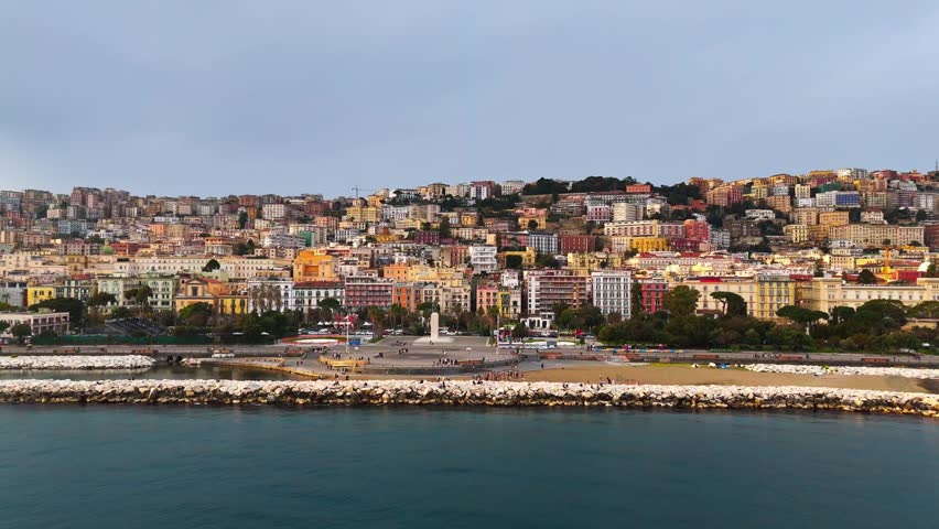 Aerial view of Naples, Italy, centered on the beach plaza, white breakwater, and densely layered hillside neighborhoods above the waterfront.