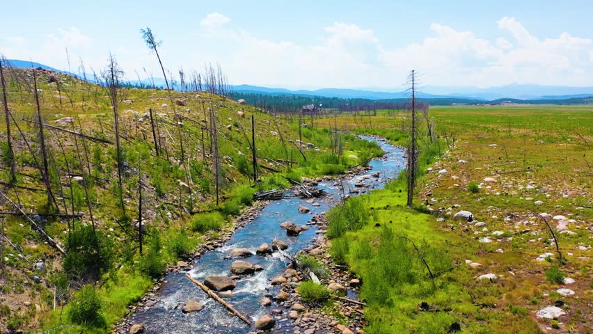 Forward flying aerial in Rocky Mountain National Park near Grand Lake Colorado following a winding stream across open meadow terrain with scattered boulders charred trees and mountain views