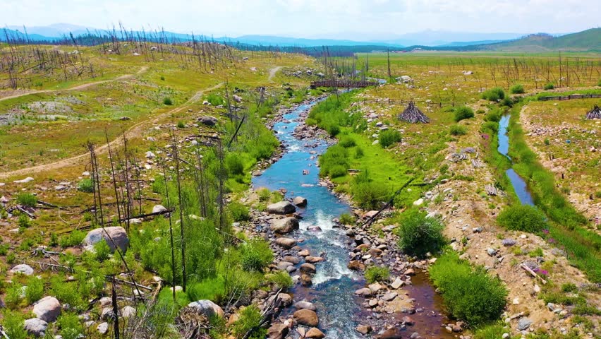 Cinematic backward moving drone view revealing a clear creek across alpine meadowland with stone lined water sparse trees sunlit grass open valley terrain and hazy peaks beyond